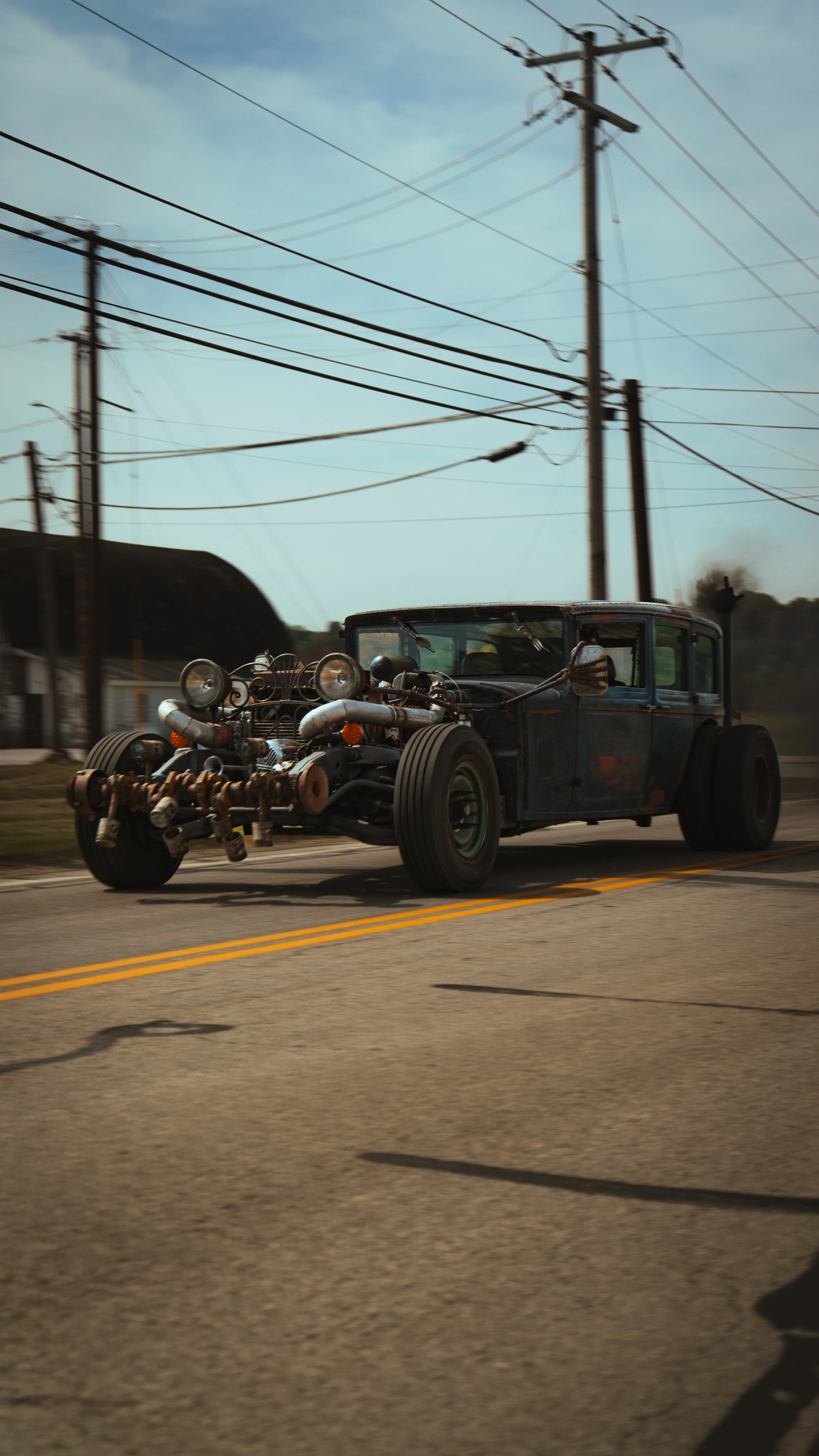 A vintage, heavily modified car with exposed engine and large tires driving on a road with power lines overhead.