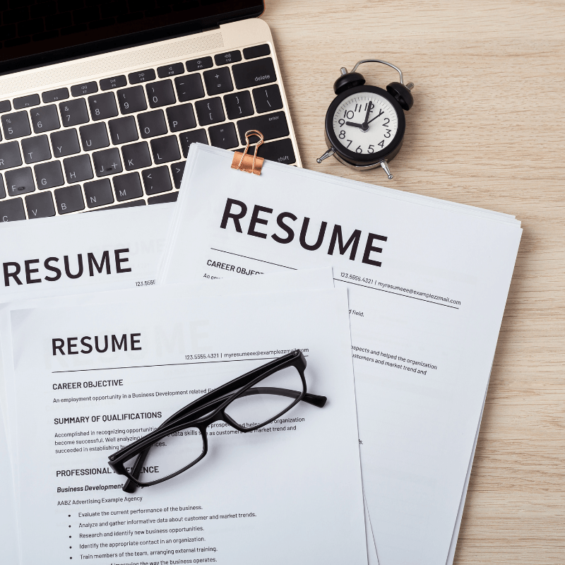 A desk with a laptop, two resumes, a pair of glasses, and an alarm clock.