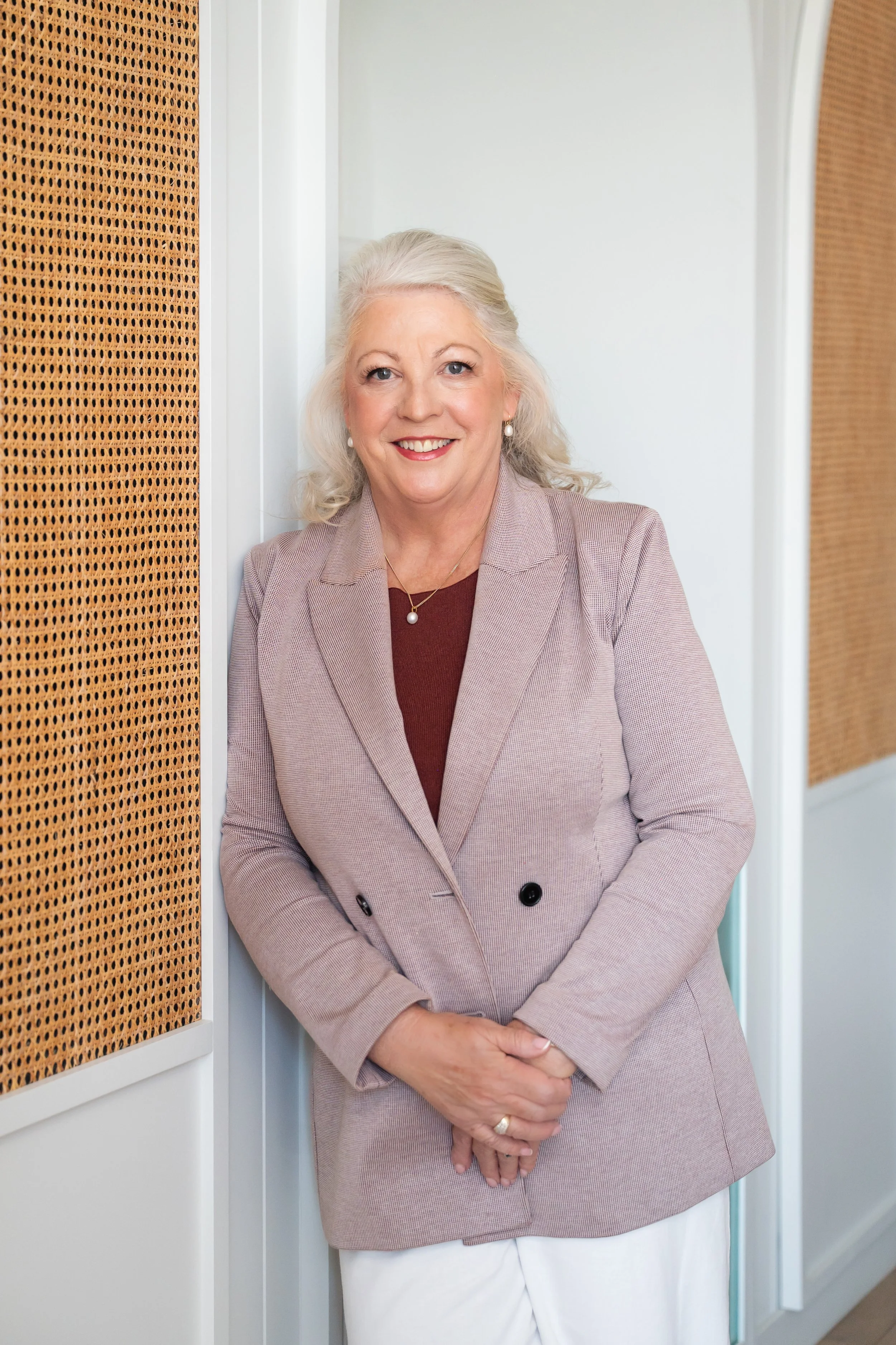 A smiling mature woman with blonde hair, wearing a tan blazer and a maroon top, standing in a modern indoor setting with white walls and wooden accents.