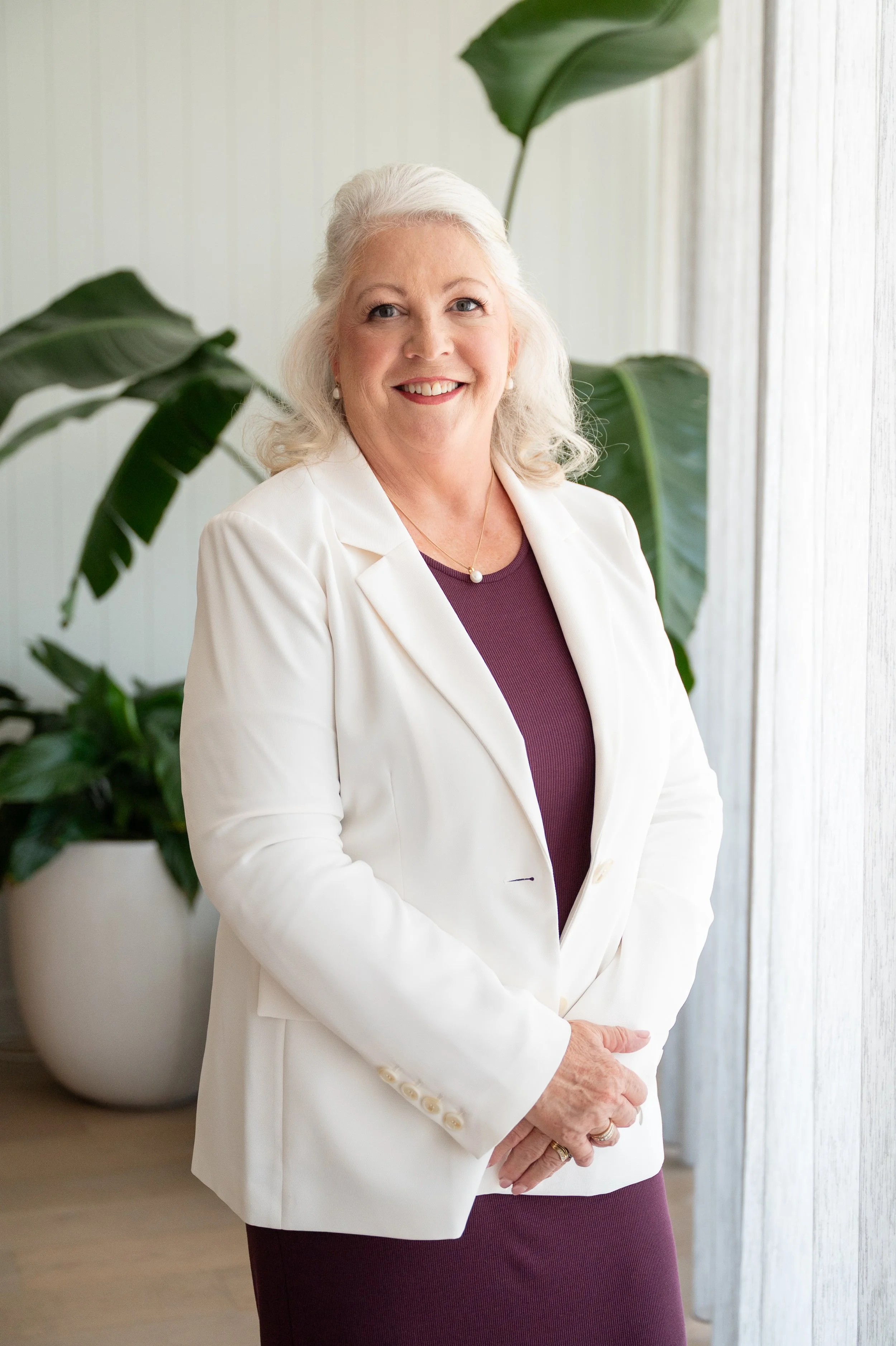 A smiling elderly woman with white hair, wearing a white blazer over a purple dress, standing indoors near a large green houseplant and sheer curtains.