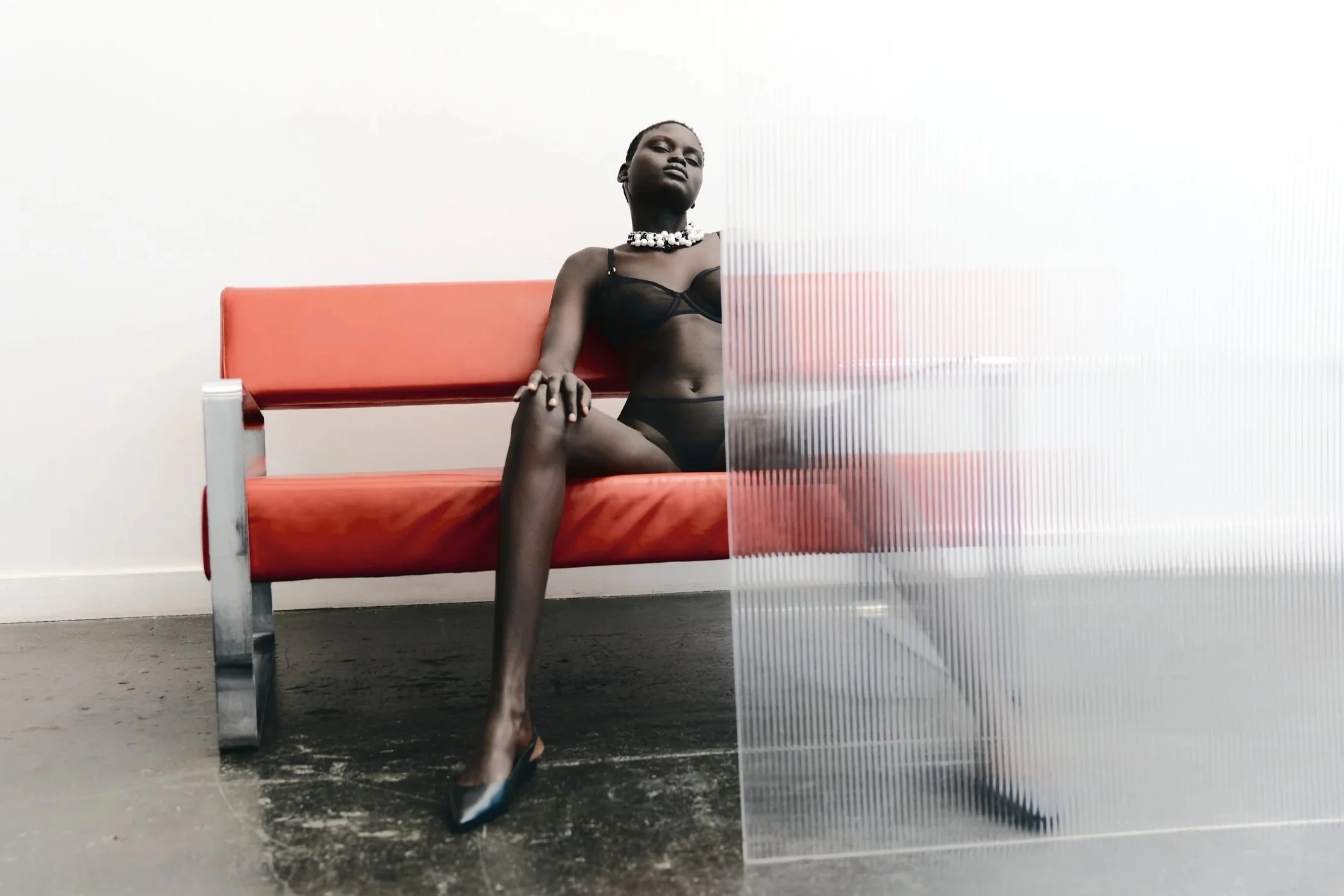 A woman wearing black lingerie, a pearl necklace, and high heels sitting on an orange bench in a room with plain white walls. A translucent, textured glass panel partially obscures her figure.