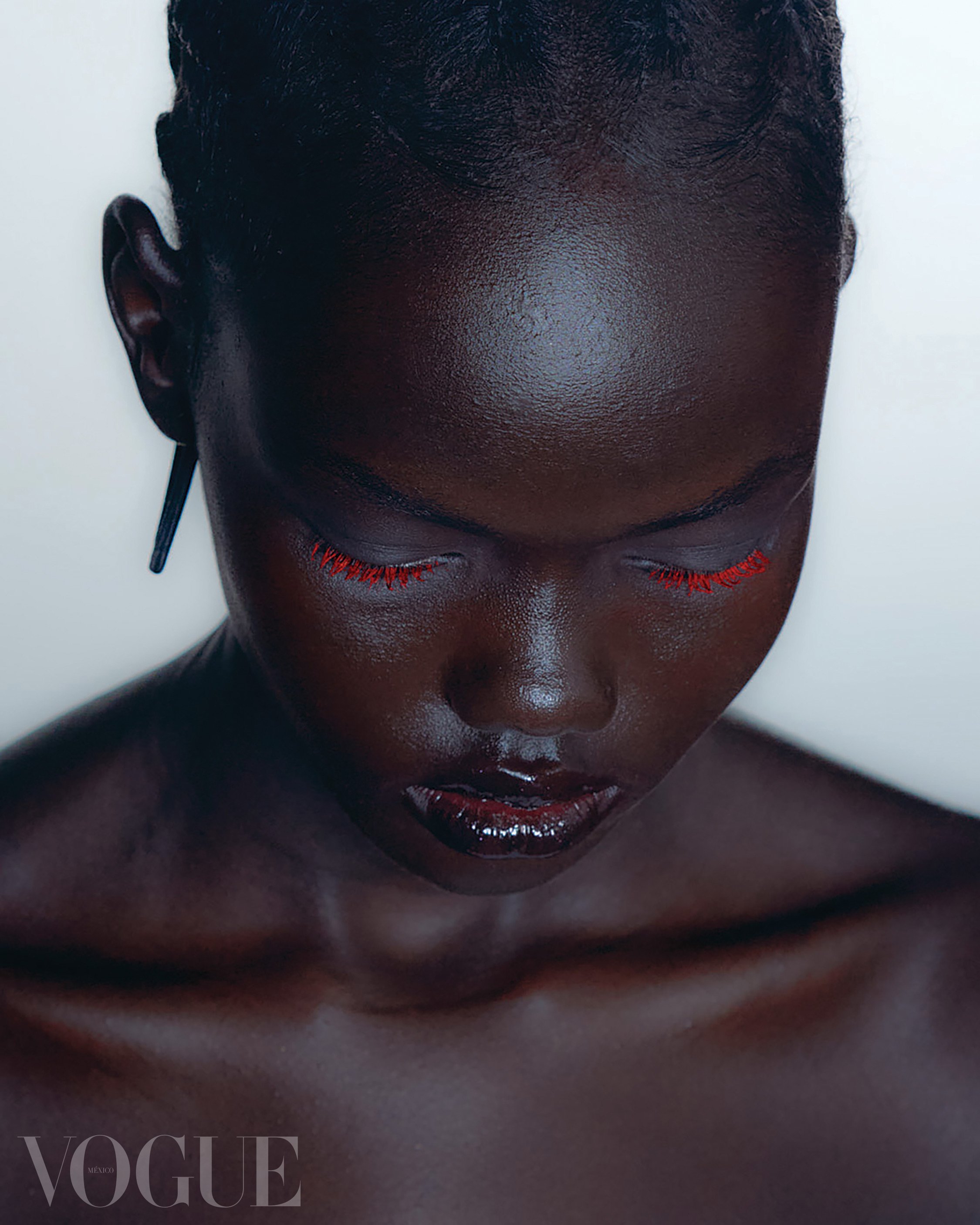 A close-up portrait of a woman with dark skin and closed eyes, wearing red eyelashes and dark lipstick, with a VOGUE logo in the bottom left corner.