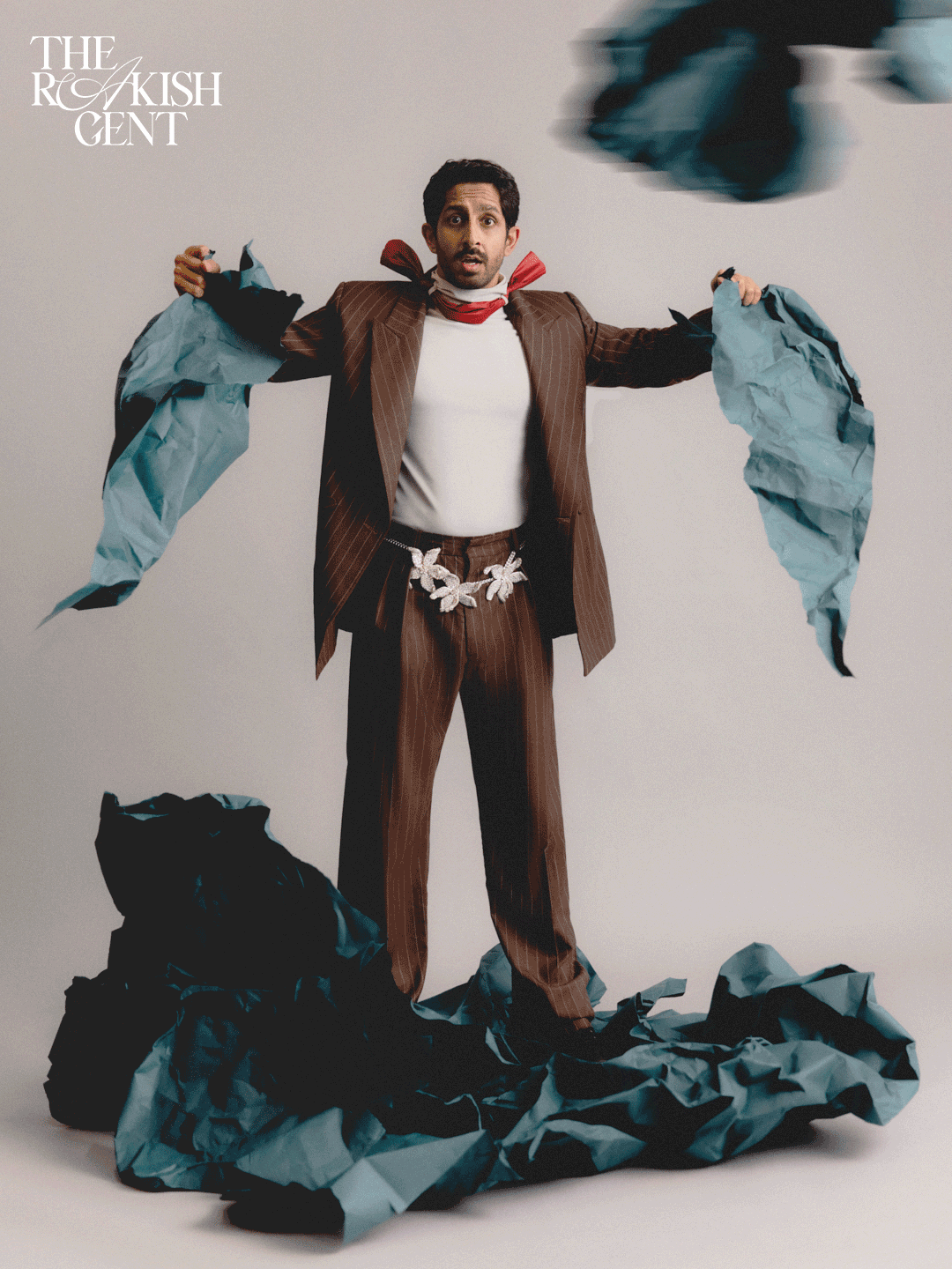 A man dressed in a brown pinstripe suit with a white shirt, red scarf, and floral belt, stands in front of a plain background surrounded by black and blue crumpled paper. He has a surprised expression, with crumpled paper in his hands and some fallin