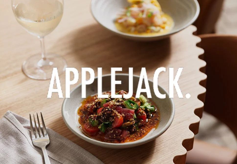 A bowl of Asian-inspired salad with tomatoes, greens, and sesame seeds on a wooden table, with a glass of white wine and another dish in the background.