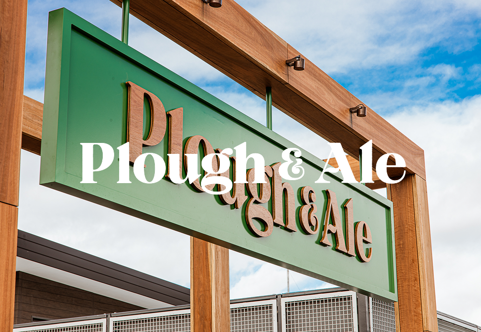 A green and brown sign for Plough & Ale, mounted on a wooden frame, with a partly cloudy sky in the background.
