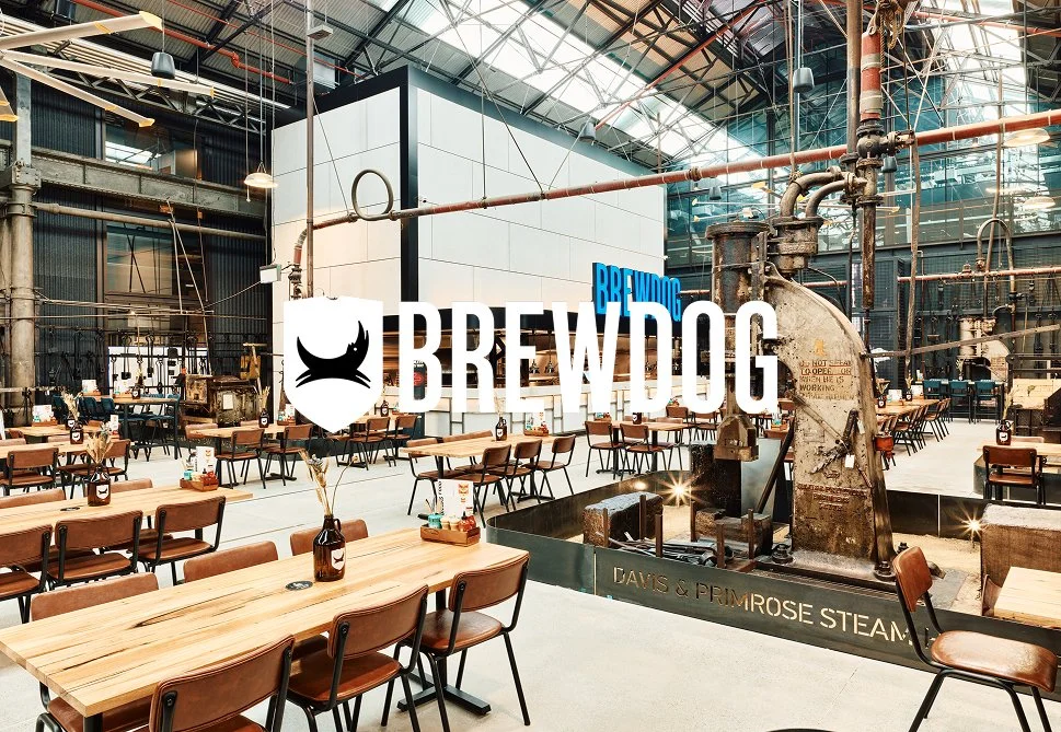 Industrial-style brewery interior with wooden tables and chairs, large machinery, and pipes. A prominent Brewdog logo is overlaid on the center of the image.
