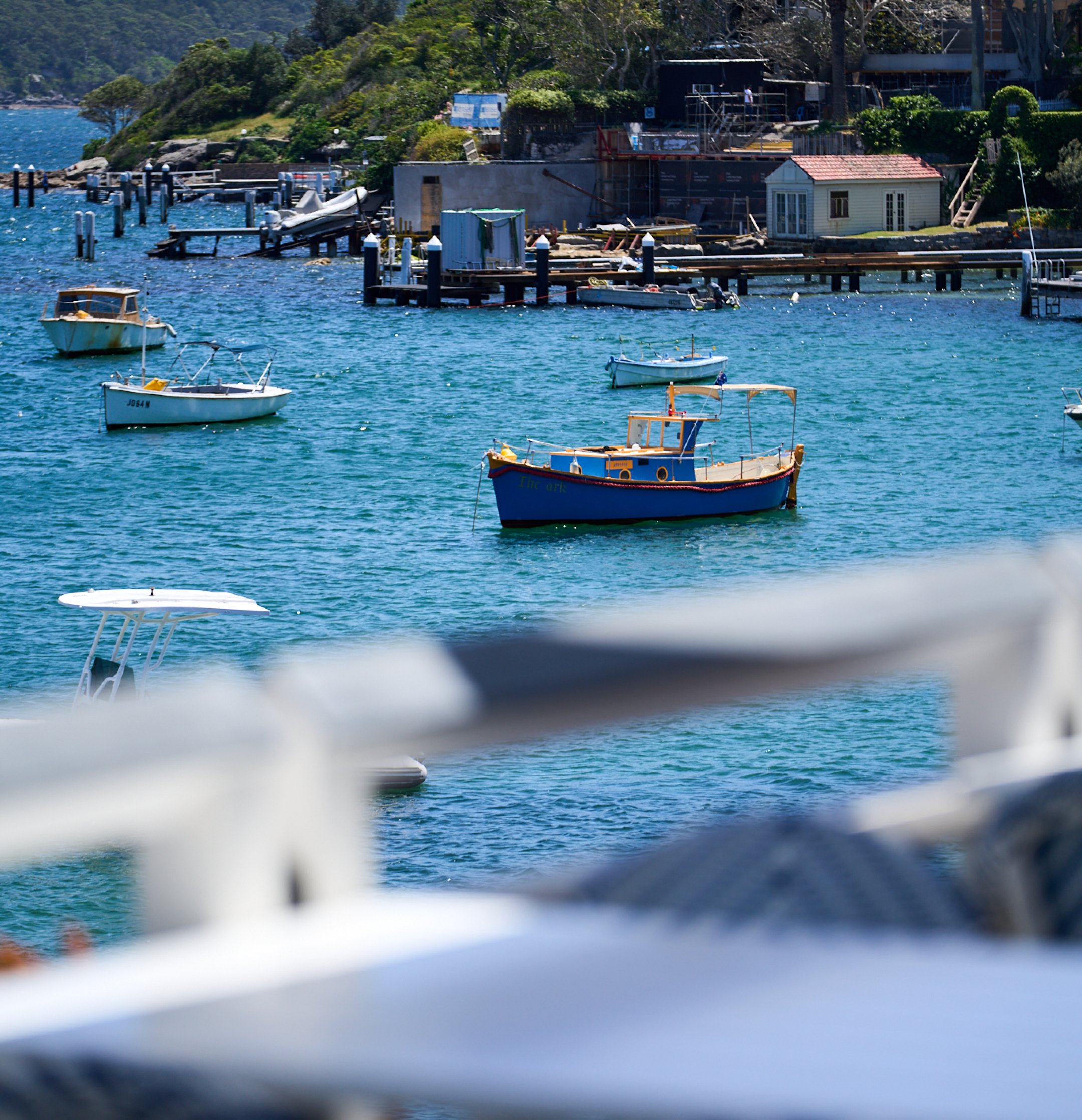 Boats anchored in a harbor with a shoreline under construction in the background, including small houses and greenery.