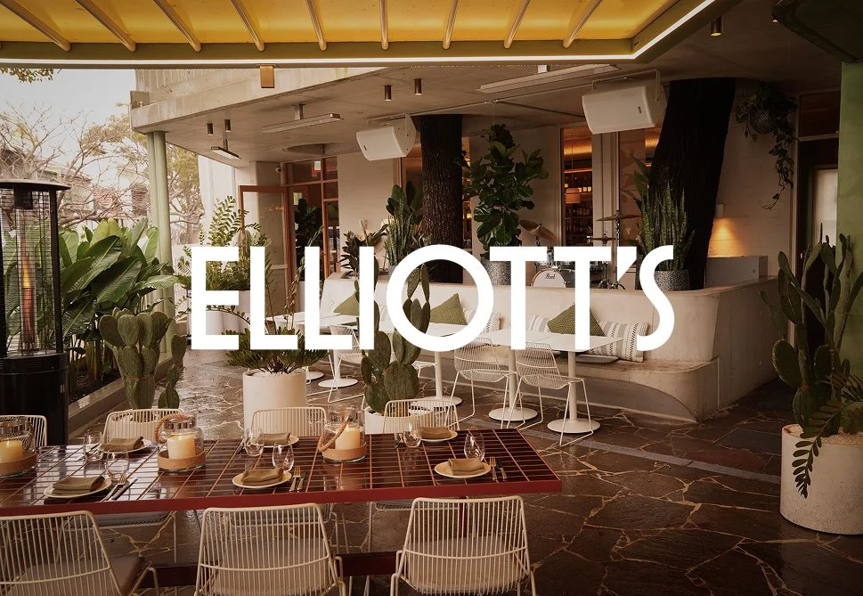 Indoor restaurant or event space with a dining table set with candles and tableware, surrounded by green plants and modern white chairs, featuring a naturalistic and stylish ambiance.