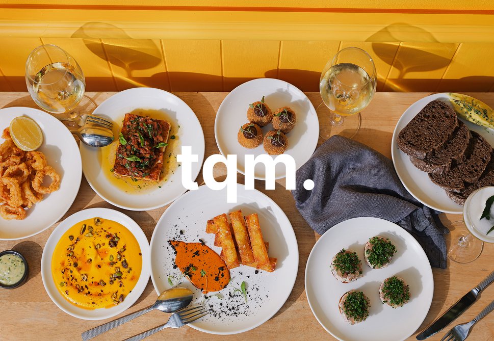 A variety of dishes on a wooden table including fried calamari, a fillet of fish with sauce, croquettes, bread slices, and small appetizers, with glasses of white wine.
