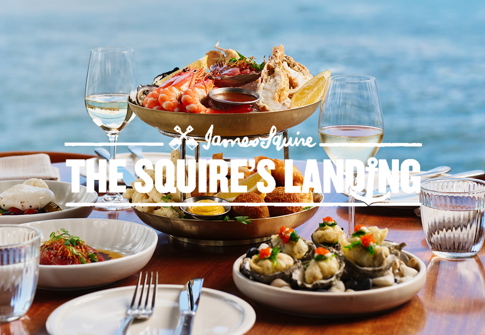 Seafood dishes on a table with a view of the ocean in the background, including a large bowl of assorted seafood, oysters topped with garnishes, shrimp, and side dishes, along with wine glasses and cutlery.