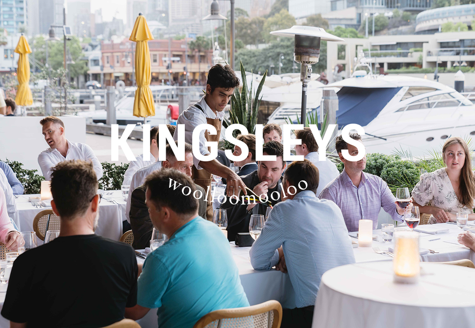 People dining outdoors at a waterfront restaurant with boats docked in the background, including a waiter serving a group of guests.
