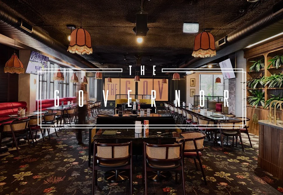 Inside a stylish restaurant with pink lampshades hanging from the ceiling, a floral carpet, and wooden accents. There are tables with chairs set for dining and a view of a garden outside.