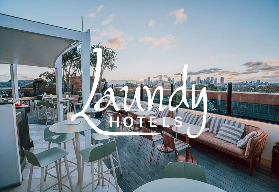 Rooftop lounge with modern furniture, outdoor seating, and city skyline view at sunset, branded with 'Laundry Hotels' logo.