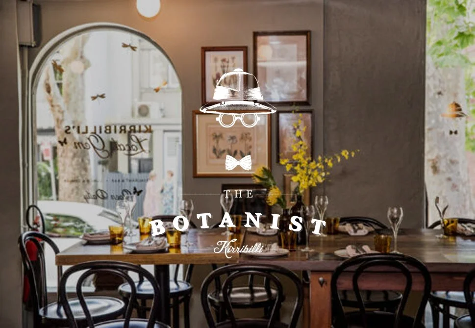 Interior of a cozy restaurant with a wooden table set with glasses and plates, yellow flowers in a vase, framed artwork on the wall, a large window with the restaurant's name, and a hanging metallic light fixture.