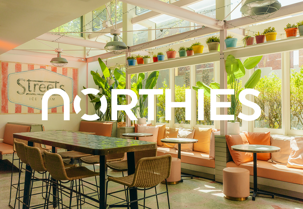 Bright, modern cafe interior with large windows, green plants, and colorful potted plants on shelves, featuring a long pink sofa, round tables, and a large sign that says 'Streets Ice Cream'