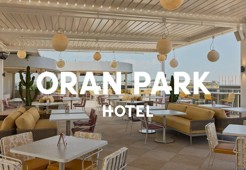 Indoor dining area at Orange Park Hotel with white tables, yellow and patterned chairs, beige sofas, tall cactus plants, hanging round light fixtures, and large open windows with a view of the city sky.