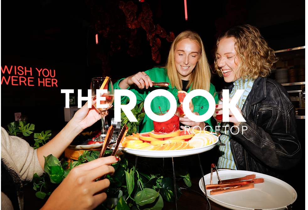 Two women celebrating a birthday with a cake, drinks, and fruit at a restaurant or bar, with a neon sign that says 'Wish you weren't her' in the background.
