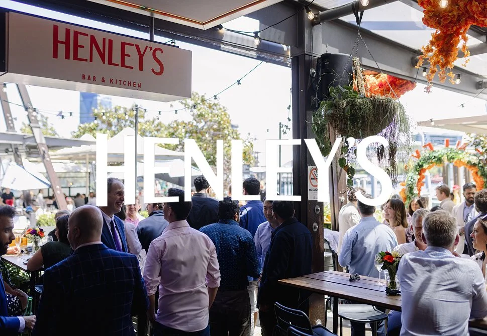 Crowd of people gathered inside and outside Henley's Bar & Kitchen, with a large Henley's sign over the entrance, some flower decorations, and outdoor umbrellas.