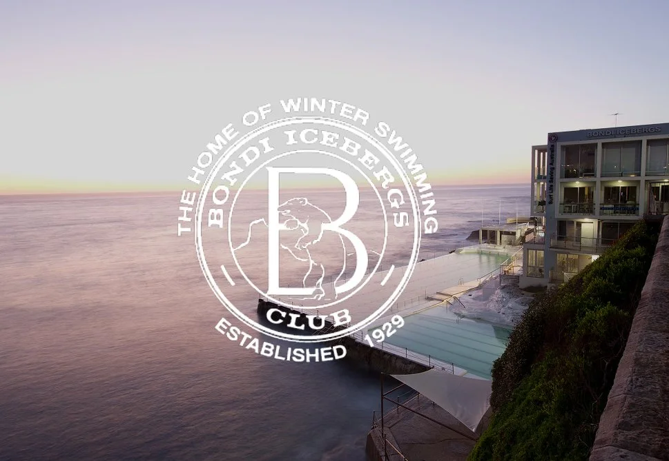 Sunset view over a coastal hotel with outdoor pools and a logo overlay for the Bondi Icebergs Club, established in 1929, featuring a large 'B' and a polar bear emblem.