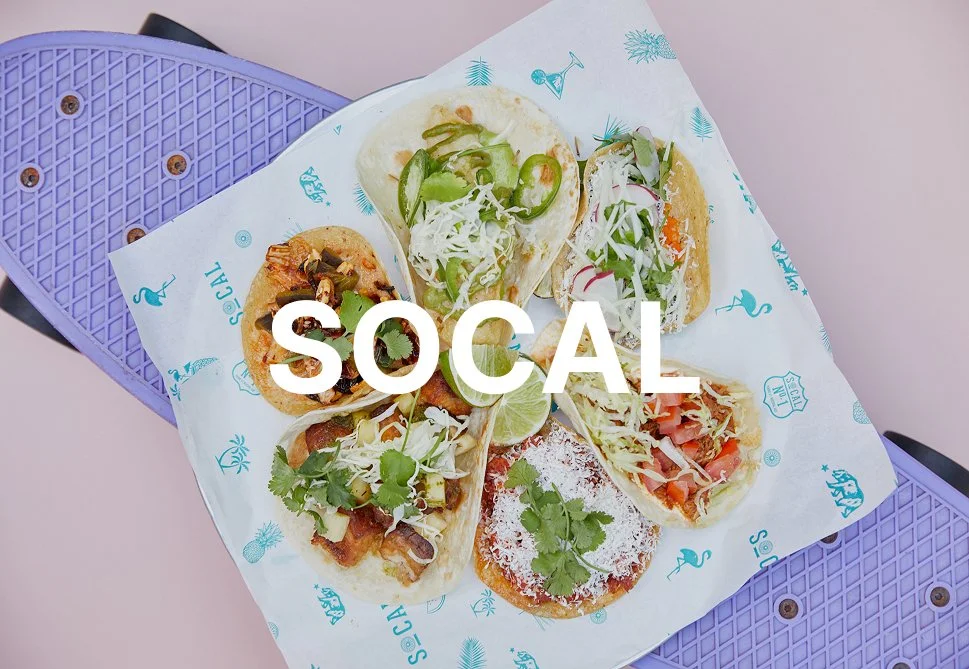A basket of six tacos with various fillings and toppings, garnished with lime wedges, on a sheet of branded paper, on a purple tray.