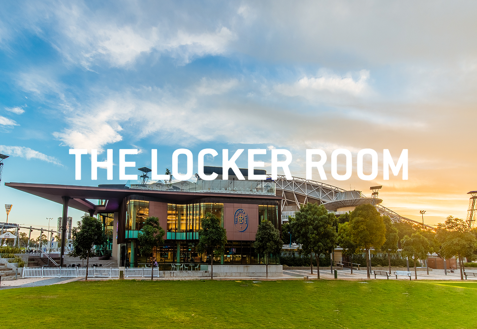 Exterior view of an arena with a modern design, trees in the foreground, and a sunset sky, with the words 'THE LOCKER ROOM' overlaid.