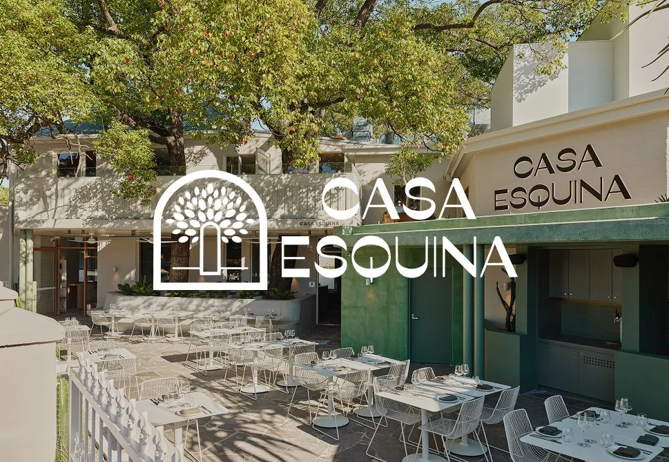Outdoor dining area with white tables and chairs, shaded by a large tree with green leaves. The restaurant, Casa Esquina, has a modern facade with signage in black lettering.