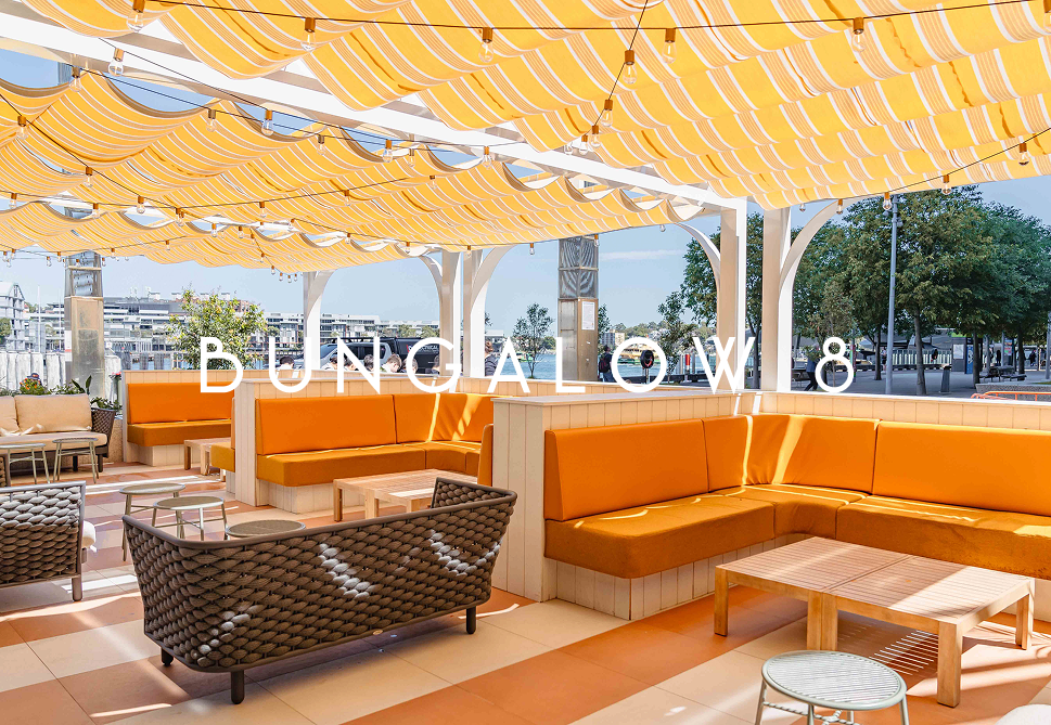 Outdoor seating area at Bunka Bow with orange cushioned benches, rattan chairs, and yellow striped awning, overlooking a waterfront with trees and buildings in the background.