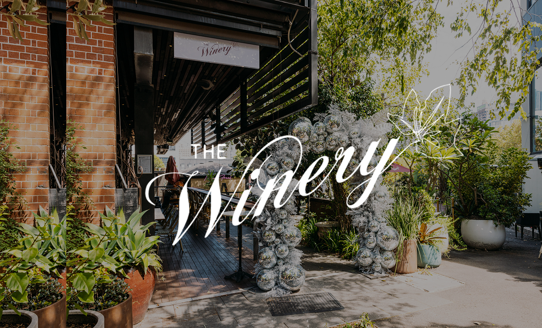 An outdoor entrance decorated with a large floral arch made of silver ornaments and greenery, leading to a wine shop or winery, with potted plants and trees around.
