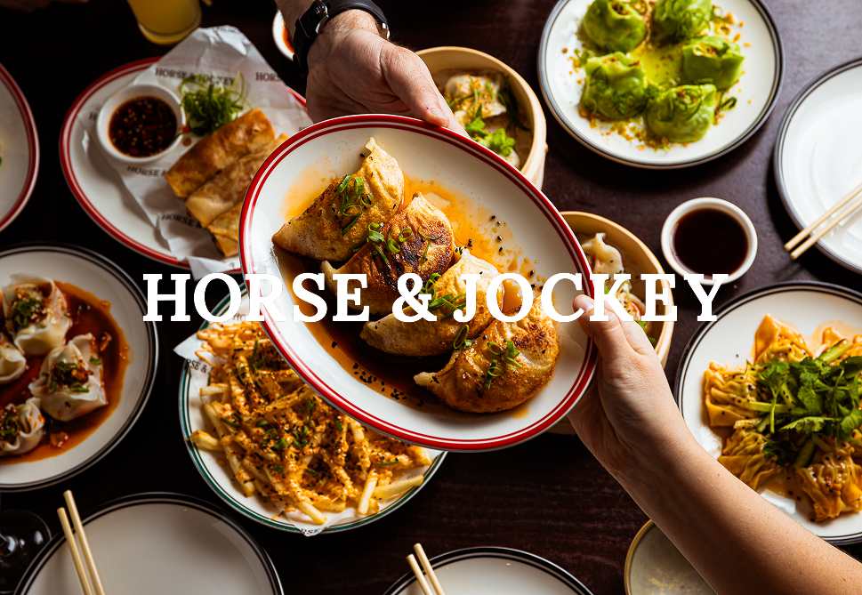 A table spread with various Asian dishes including dim sum, dumplings, and spring rolls, with a person holding a plate of fried spring rolls garnished with green onions.