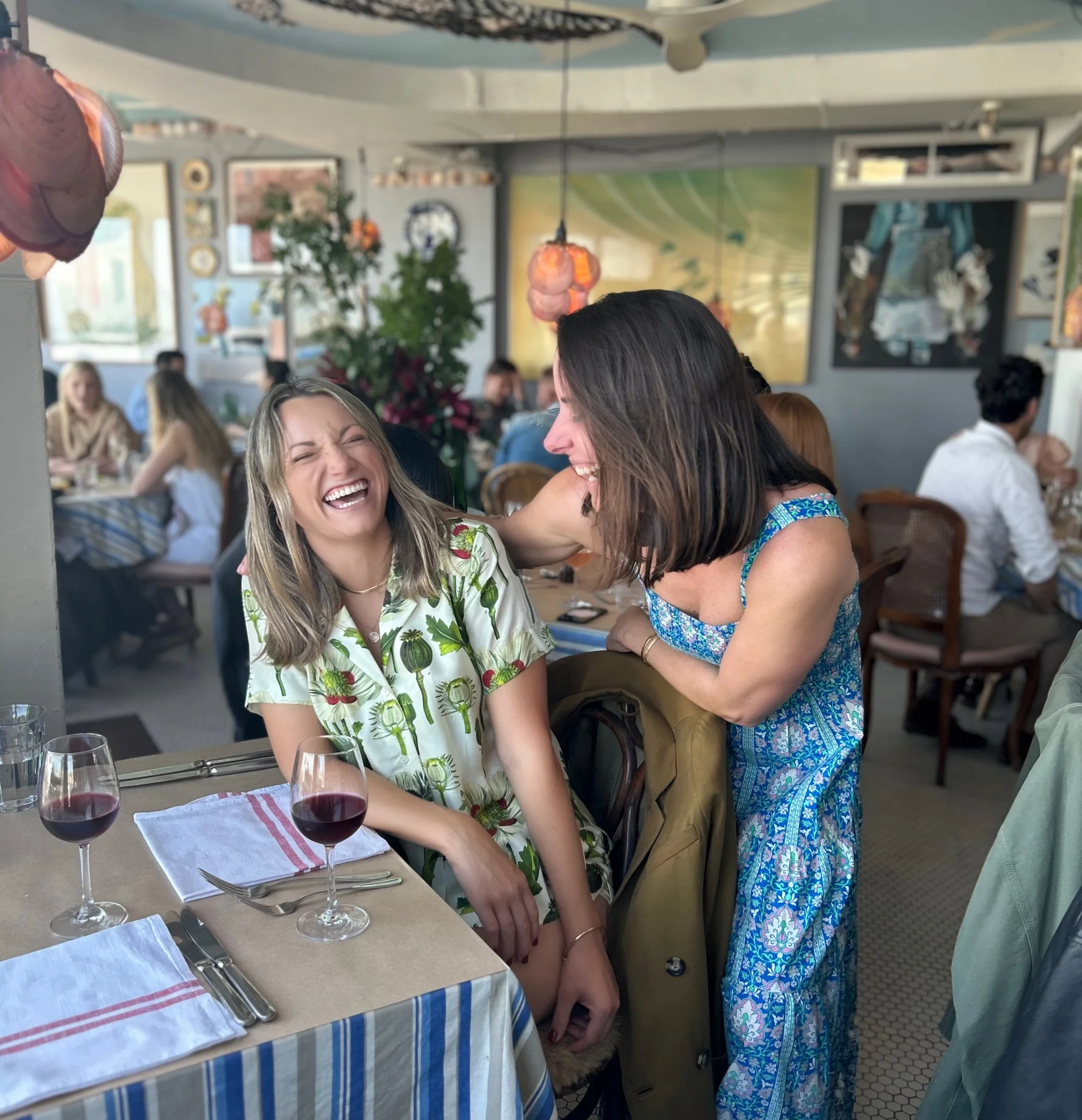 Two women are laughing and smiling at a restaurant table. One woman with straight brown hair wearing a blue floral dress is leaning toward a woman with blonde hair wearing a white dress with green plants. There are glasses of red wine and dinnerware on the table.