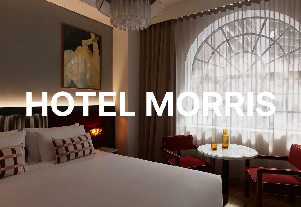 Hotel room with a bed, artwork, round table with two red chairs, and large arched window with sheer curtains.