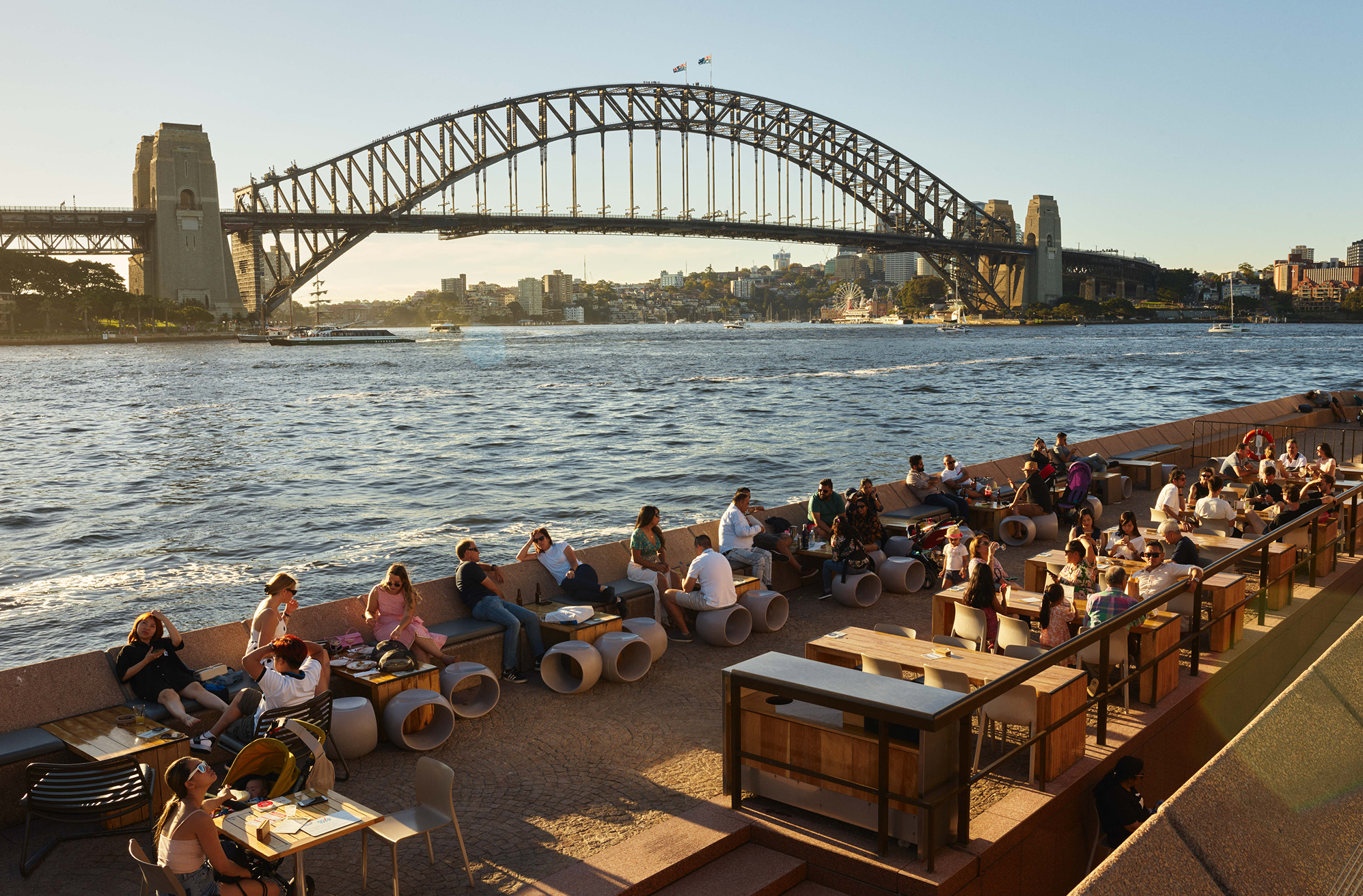 People sitting and dining outdoors near the water with the Sydney Harbour Bridge in the background during sunset.
