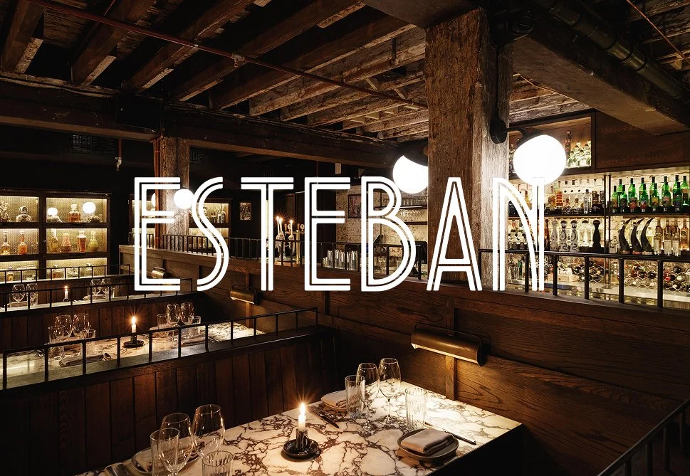 Interior of a dimly lit restaurant with wooden beams and shelves, set tables with candles, and neon sign reading 'Esterban'.