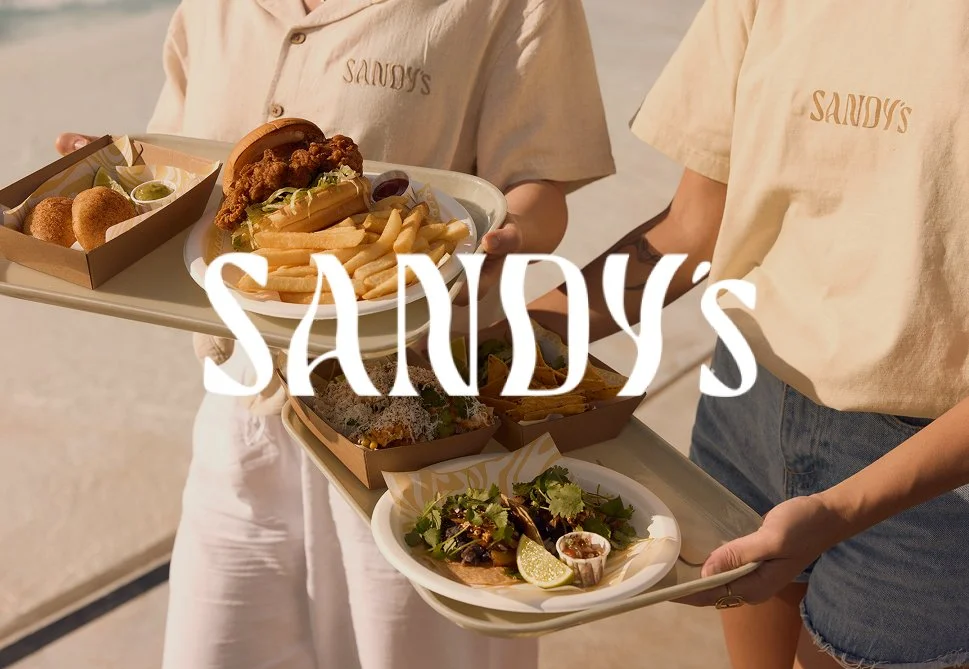 Two people wearing beige shirts with 'SANDY'S' embroidered on them, each holding a tray of food. The trays contain fried items, French fries, a sandwich, and a salad with lime wedges.