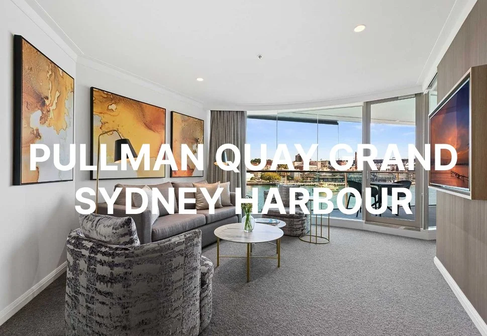 Living room in a modern apartment with grey furniture, abstract artwork on white walls, a large window with city view, and a flat-screen TV.