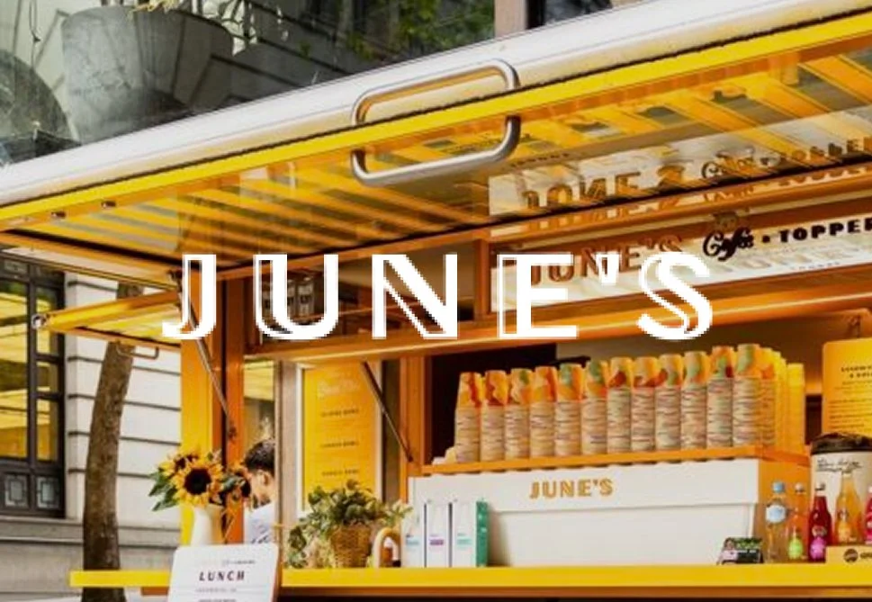 Yellow and orange street food cart named 'June's' selling rolled sandwiches and drinks, with a sunflower arrangement and a person in the background.