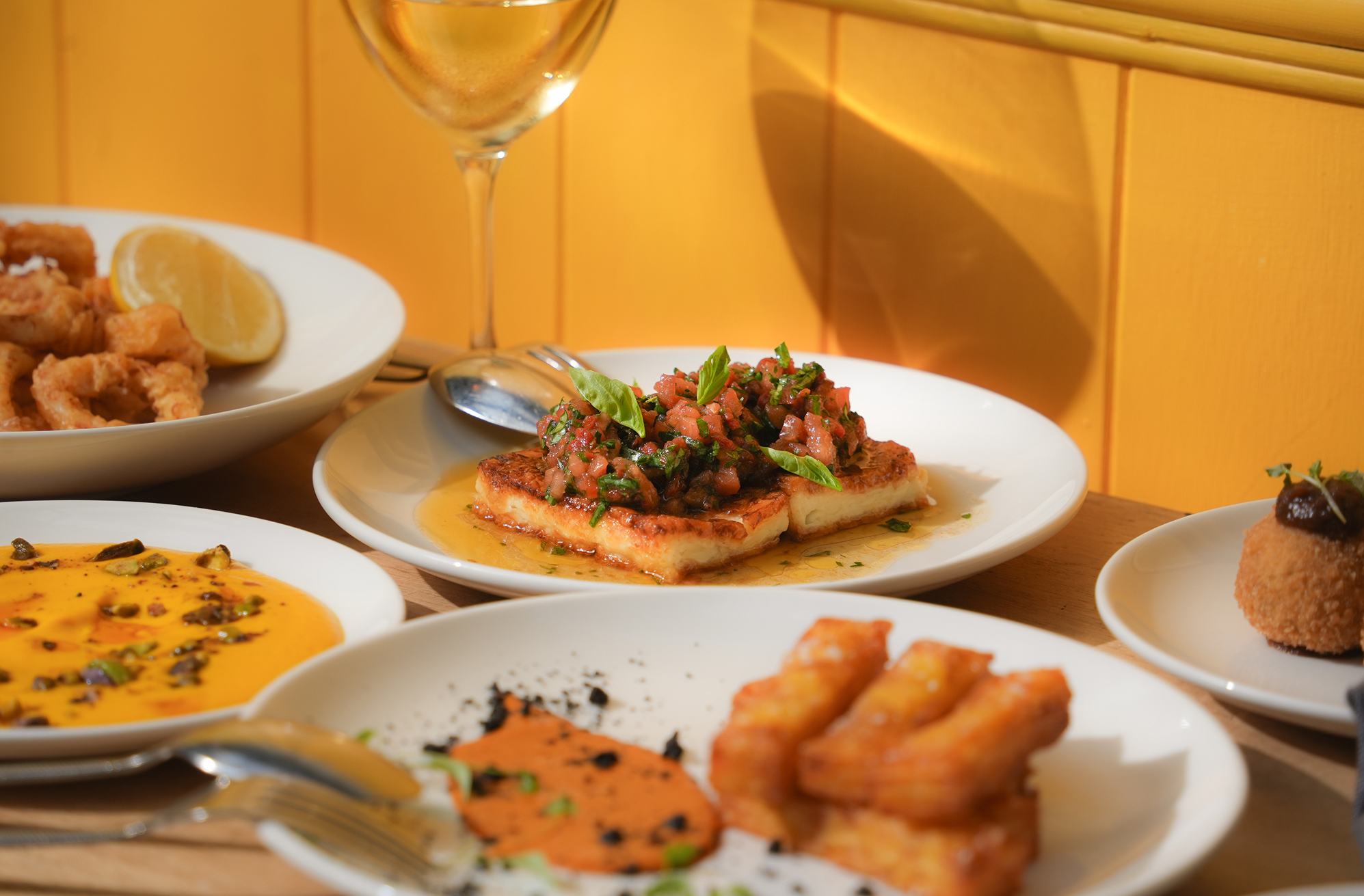 Assorted dishes of Mediterranean or Middle Eastern cuisine, including fish topped with pico de gallo salsa, fried calamari with lemon, chickpea or lentil soup, fried cheese sticks, and croquettes on various white plates on a wooden table against a yellow wall.
