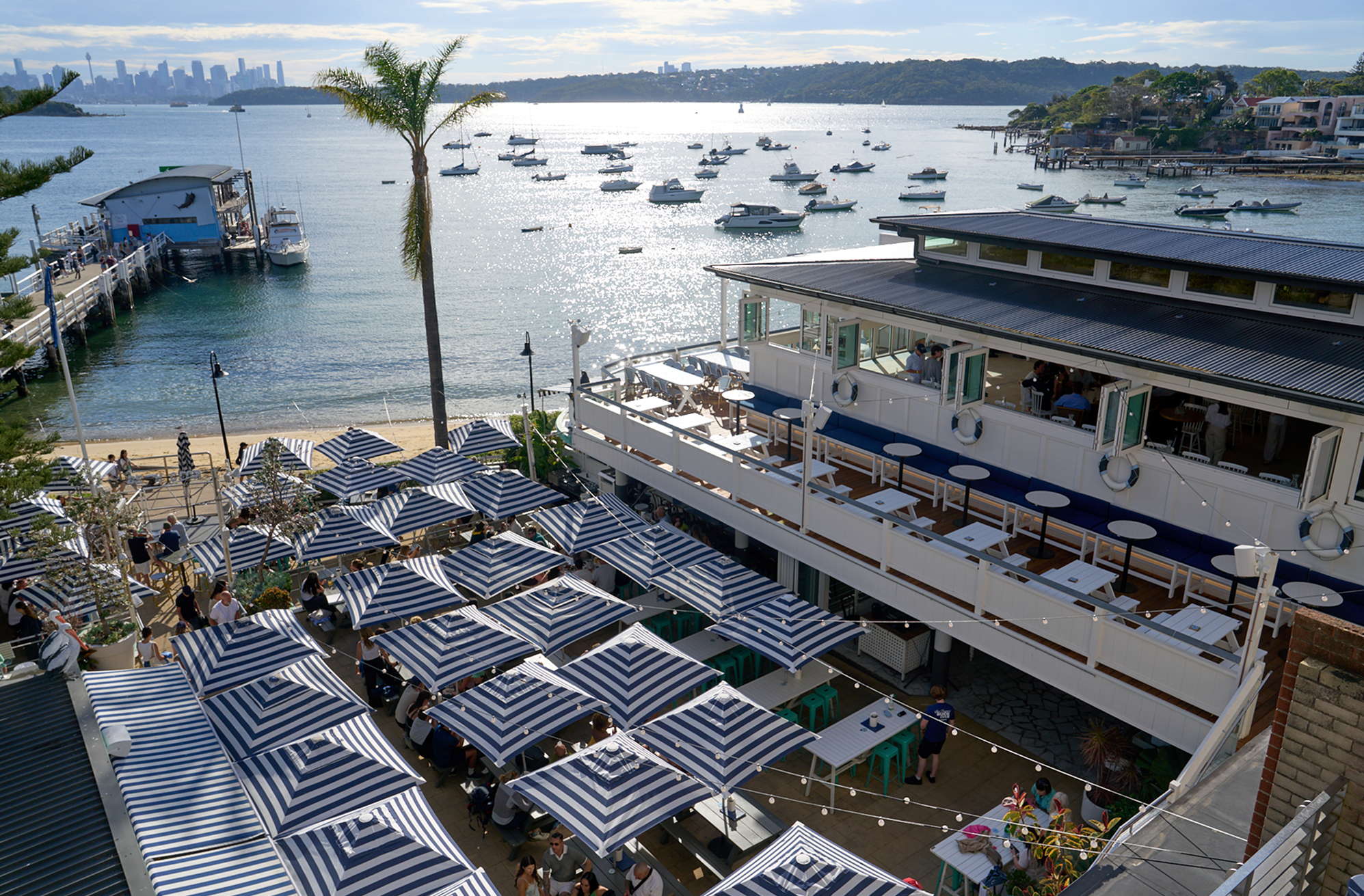 An outdoor waterfront restaurant with seating areas shade by blue and white striped umbrellas, overlooking a pier with boats and a body of water, with city skyline and green hills in the background.