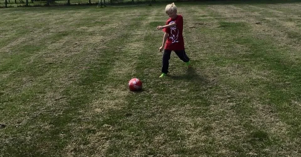 A young boy wearing a red shirt and dark pants kicking a red and white soccer ball on a grassy field.
