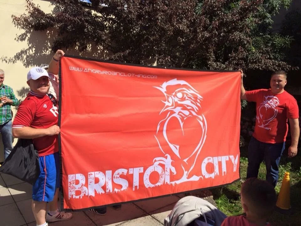 Two men holding a large red Bristol City flag outdoors, with a tree and some children nearby.