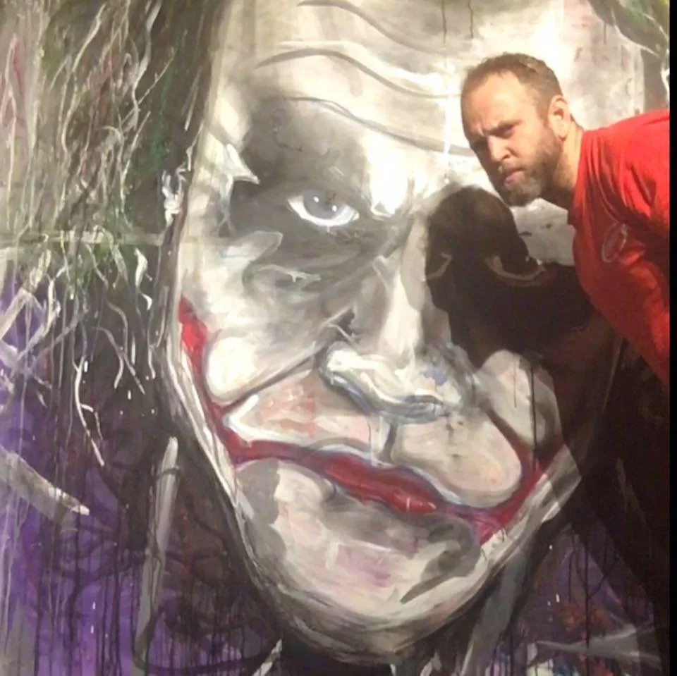 A man with a beard in a red shirt posing next to a large mural of a woman's face with clown makeup, including red lips and white face paint.