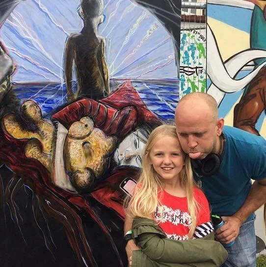 A man and young girl in front of colorful graffiti art on a wall. The girl is smiling, and the man is leaning toward her with his tongue slightly out.