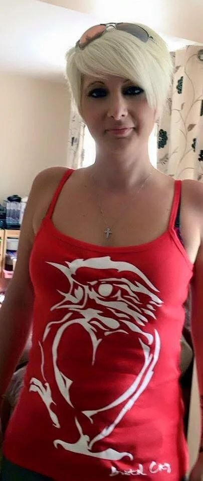 Woman with blonde hair wearing a red tank top with a white dragon and heart design, glasses on head, in a room with beige curtains and a bookshelf.