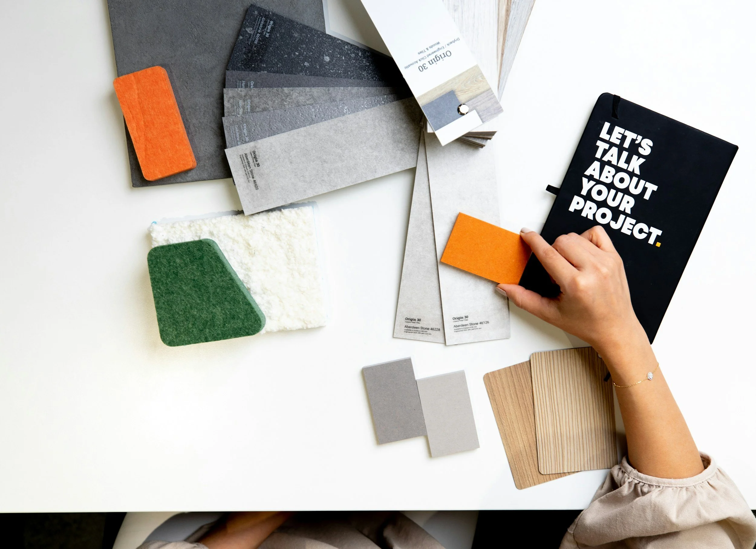 Eine Hand berührt eine orangefarbene Texturprobe auf einem Tisch. Daneben liegen Musterkarten für Bodenbeläge in Grautönen, ein Stoffmuster, eine Holzprobe und ein schwarzes Notizbuch mit weißen Buchstaben, die sagen 'Let's talk about your project'. Das Bild zeigt eine kreative Arbeitsumgebung für Innenraumgestaltung.