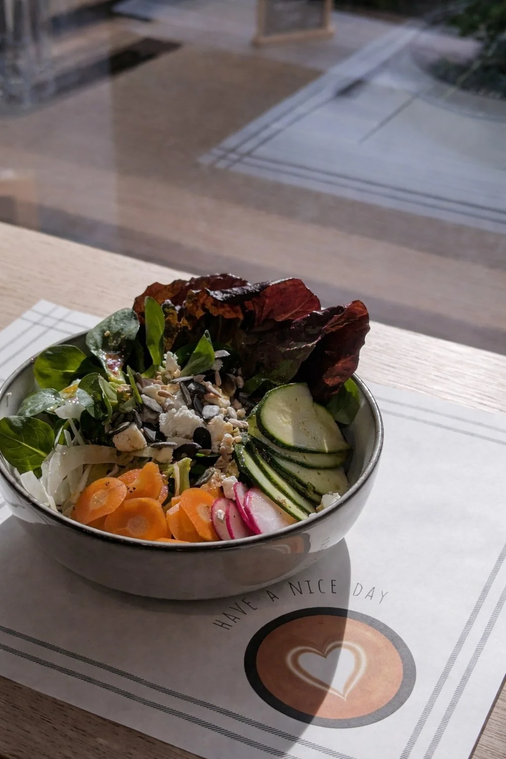 Schüssel mit gemischtem Salat, einschließlich Salatblätter, Gurkenscheiben, Radieschen, Karotten und Sonnenblumenkerne, auf einem Tisch.