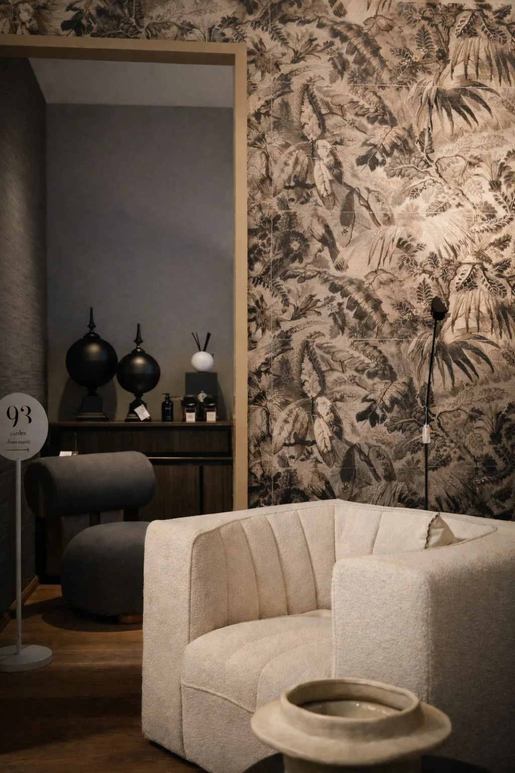 Ein stilvoll eingerichtetes Wohnzimmer mit einer beige Couch, einer Stehlampe, einer Wand mit tropischem Blattmuster, und einer dunklen Regalwand mit Dekorationsgegenständen.