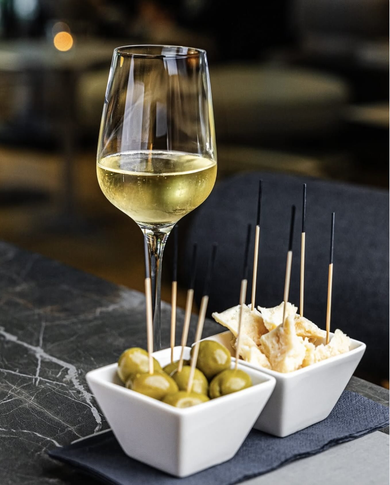 Ein Weinglas mit weißem Wein, zwei kleine Schälchen mit grünen Oliven und Käse, serviert auf einem dunklen Marmortisch.