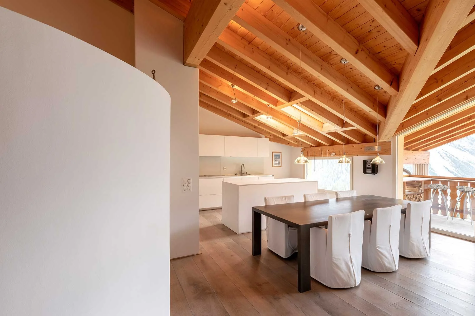 Chalet Innenansicht einer modernen Küche und Essecke mit Holzdecke, weißen Möbeln und einem großen Esstisch mit weißen Stühlen, Blick auf einen Balkon mit Bergblick.