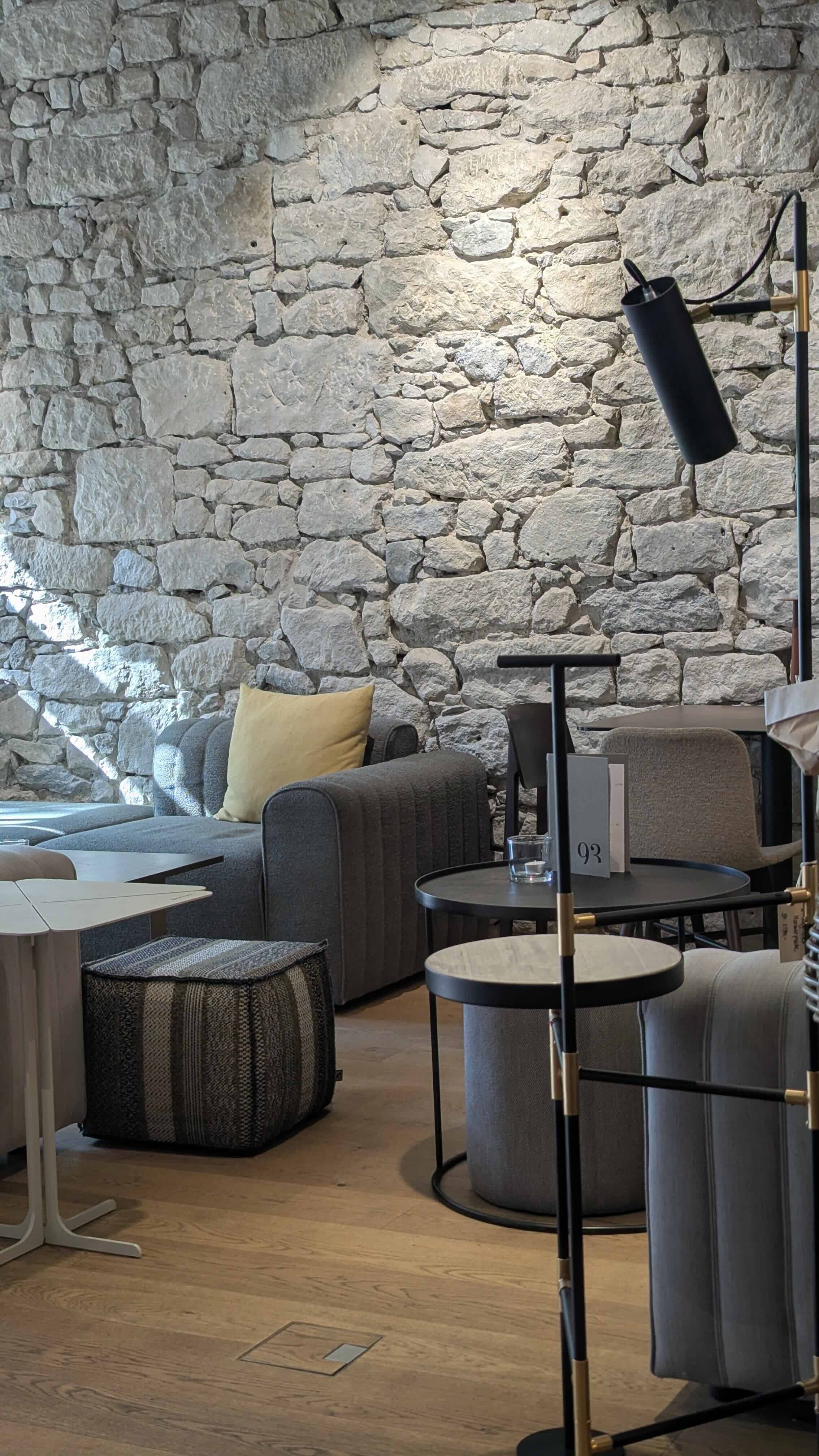 Innenraum eines modernen Wohn- oder Lounge-Bereichs mit einer hellen, steinernen Backsteinwand, dunklem Sofa mit gelbem Kissen, mehreren kleinen Tischen, einem Steh- oder Stehlampensystem und Holzfußboden.