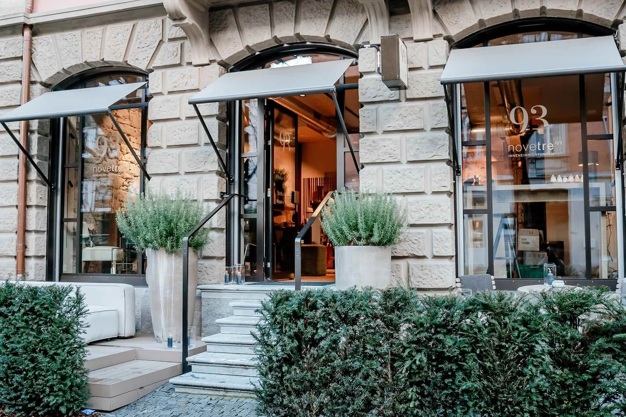 Außenansicht eines Cafés oder Restaurants mit zwei großen Fenstern, einer Eingangstür, Blumentöpfen, kleinen Tischen, Stühlen und Pflanzen im Vordergrund